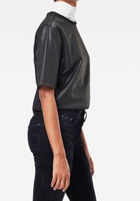 Person som bär en svart kortärmad lädertröja över en vit polotröja och svarta åtsittande jeans, visad från sidan mot en vit bakgrund.