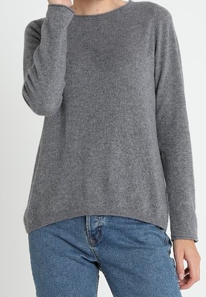 Personne portant un pull gris à manches longues avec un col rond et un jean bleu, la main couvrant partiellement le visage.