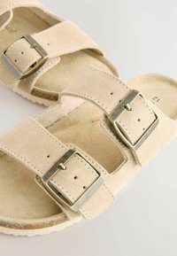 Helle Wildledersandalen mit zwei verstellbaren Riemen, silbernen Metallschnallen und einem gepolsterten Fußbett. Strukturierte Laufsohle für guten Halt und Langlebigkeit.