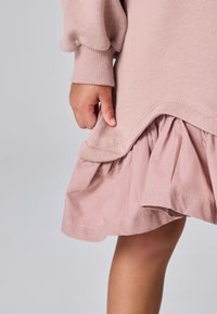 Lapsen käsi pitää vaaleanpunaisen neuleenreunan reunaa, joka sopii röyhelöiseen hameeseen, paljas polvi näkyy yksiväristä taustaa vasten.