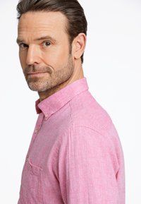 Vaaleanpunainen napillinen paita, joka on valmistettu tekstuurillisesta kankaasta. Paidassa on kaulus, rintatasku ja kaksi nappia hihassa.