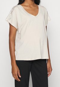 Beige top s výstřihem do V a krátkým rukávem, který má krajkové akcenty na ramenou, vyrobený z měkké tkaniny, v kombinaci s černými kalhotami.