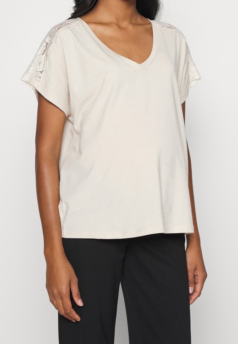 Beige top s výstřihem do V a krátkým rukávem, který má krajkové akcenty na ramenou, vyrobený z měkké tkaniny, v kombinaci s černými kalhotami.