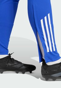 Blaue Sporthosen mit seitlichen Reißverschlüssen und drei weißen vertikalen Streifen. Getragen mit schwarzen Fußballschuhen mit strukturiertem Design.