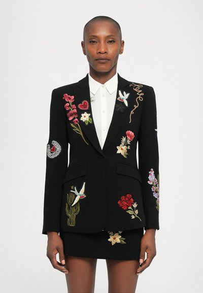 Person iført en sort skræddersyet blazer og nederdel med farverig blomster- og fuglebroderi over en hvid skjorte med knapper, stående foran en hvid baggrund.
