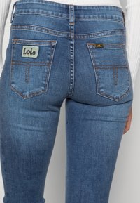 Tunna blå denimjeans med en ljusblå logotyp på den högra bakfickan och gyllene detaljer på den vänstra fickan.