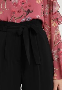 Pantalones negros de cintura alta con un cinturón atado, combinados con una blusa rosa con flores que tiene mangas con volantes y un tejido texturizado.