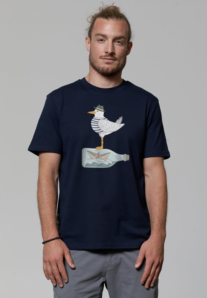 Navyblauwe katoenen t-shirt met een afbeelding van een meeuw in een gestreept shirt en hoed, staande op een fles met een papieren bootje erin.