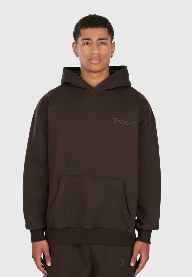 Giovane uomo che indossa una felpa a cappuccio marrone scuro con tasca frontale e logo "Dropsize" sul petto, in piedi contro uno sfondo chiaro e uniforme.