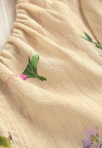 Tejido ligero con un patrón texturizado y tejido; presenta diseños florales en rosa y verde, destacándose sobre un suave fondo beige. Acentos de brillo sutil.