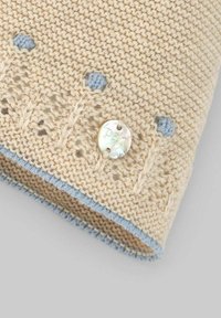Cappello beige lavorato a maglia con rifinitura azzurro chiaro, caratterizzato da motivi testurizzati, piccole pom pom blu e un bottone perlaceo cucito di lato.