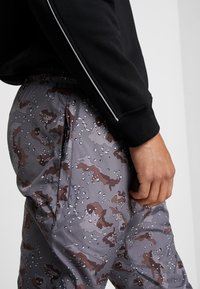 Pelēkie kamuflāžas bikses ar brūniem akcentiem, ar sānu kabatu ar rāvējslēdzēju un elastīgu jostasvietu. Materiāls šķiet viegls.