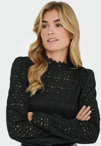 Juodas megztas megztinis su tekstūruotu raštu, turintis aukštą nėriniuotu apykaklę ir ilgomis pūstomis rankovėmis.