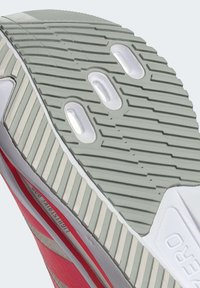 Podešev obuvi s šedou gumou, texturovanými drážkami a třemi ventilačními otvory, zvýrazněná bílým okrajem a červeným bočním detailem.