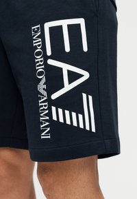 Tmavomodré bavlnené šortky s veľkým bielym logom "EA" a nápisom "Emporio Armani" na boku, pod logom so zvislým pruhom.