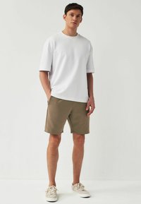 Chemise blanche à manches courtes avec un col rond, associée à un short vert olive longueur genou. Des baskets beiges complètent la tenue.
