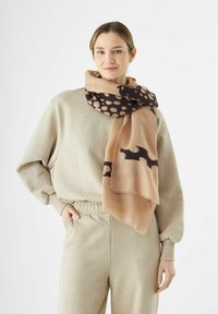 Écharpe beige avec un motif tacheté marron foncé, fabriquée dans un tissu doux et texturé, enroulée autour du cou, complétée par une tenue gris clair.