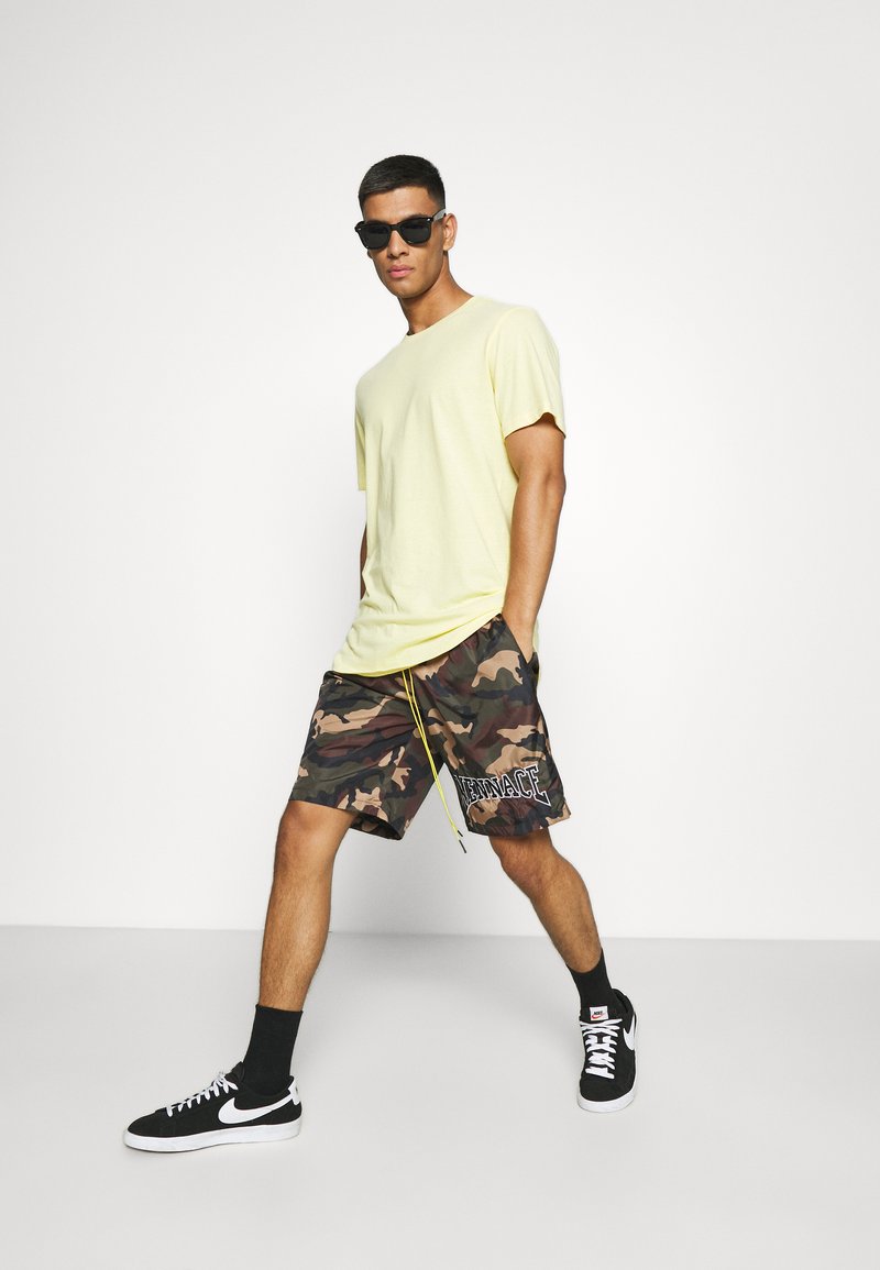 Gelbes Baumwoll-T-Shirt kombiniert mit Camouflage-Shorts mit Kordelzug und "VENICE"-Textverzierung. Schwarze Sneakers und Sonnenbrille inklusive.