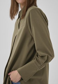 Frau in einem olivgrünen, knopfverschlossenen Hemd mit langen Ärmeln, teilweise mit der Hand in der Tasche einer blauen Jeans, vor einem schlichten hellen Hintergrund.