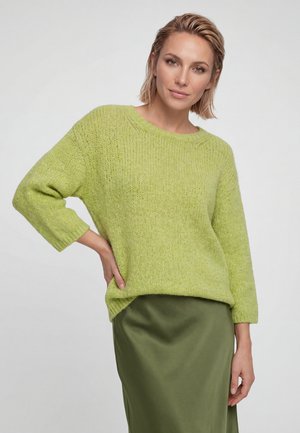 Femme aux cheveux blonds courts portant un pull en maille vert clair et une jupe en satin vert foncé, posant avec une main sur la hanche.