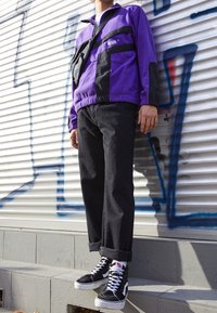 Lilla og sort fleecejakke med lynlås i nakken, forlommer og elastisk talje; sorte jeans med oprullede manchetter; sorte højhælede sneakers.
