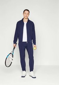 Námornícka športová bunda so zipsom vpredu, v kombinácii s priliehavými námorníckymi nohavicami, bielym topom, drží tenisovú raketu a loptičku. Hladká textúra látky.