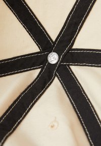 Tela crema con costuras negras que forman un patrón en X, con un botón plateado grabado con "47". El diseño enfatiza la textura y el contraste.