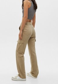 Vrouw in beige cargobroek, witte sneakers en een grijze mouwloze crop top staat zijwaarts tegen een effen achtergrond.