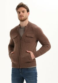 Cardigan a maglia marrone con chiusura zip, dettagli a coste e due tasche sul petto. Indossato sopra una camicia grigia chiara. Design casual fit.