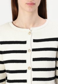 Strikkegenser i krem med sorte striper, med rund hals og dekorative gullknapper ved lukningen.