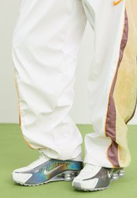 Witte sportbroeken met oranje en bruine accenten, gecombineerd met zilverkleurige en witte sneakers met een reflecterend, iriserend ontwerp en zichtbare luchtkussens.