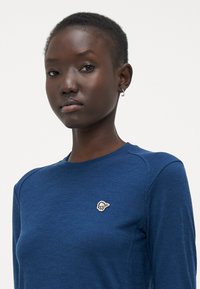 Long-sleeve navy blue top made from textured fabric, featuring a small white logo on the left chest and subtle shoulder seams.