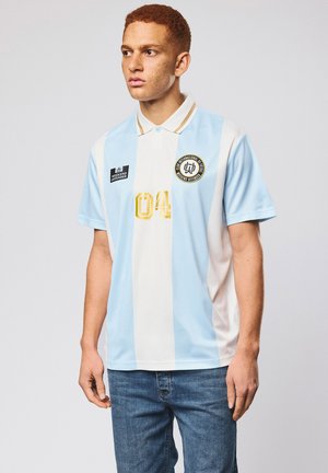 Młody mężczyzna z rudymi włosami, ubrany w jasnoniebieską i białą koszulkę polo w pionowe paski z logo i złotym numerem 04, stojący na jednolitym tle.