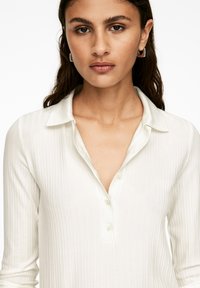 Young woman with long dark hair wears silver hoop earrings and a white ribbed long-sleeve shirt with collar and buttons.