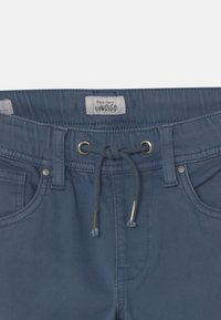Pantaloni in denim azzurro chiaro con vita regolabile tramite cordoncino, accessori in metallo, passanti per cintura e tasche frontali. L'etichetta reca la dicitura "Pepe Jeans GYWDIGO."