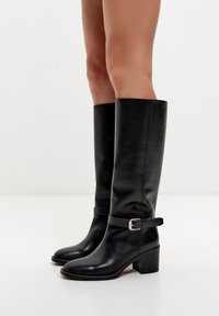 Mustat nahkaiset polven yli ulottuvat saappaat, joissa pyöreä kärki, hopeinen solkikoriste nilkkahihnassa ja paksu, pinottu korko. Sileä pinta.