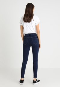 Jeans ajustados de tiro alto en denim elástico de color azul oscuro, con bolsillos traseros, combinados con una camiseta blanca y zapatillas negras.