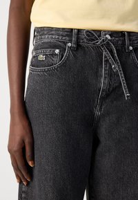Sorte denimbukser med snor i midjen, lys søm, og en liten grønn Lacoste-logo på framlommen. Myk tekstur og avslappet passform.
