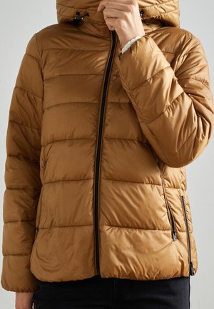 Personne portant une doudoune marron à capuche, partiellement zippée, ajustant le curseur de la fermeture éclair près du cou.