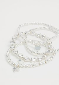 Štiri srebrne in biserne zapestnice z obeski v obliki srca in perlicami, razporejene tako, da se prekrivajo na beli površini.