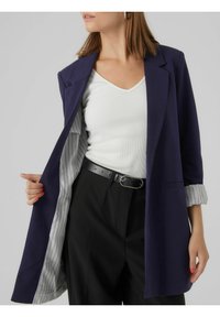 Marineblauw colbert met een getailleerde pasvorm, voorzien van een lichte gestreepte voering. Gecombineerd met een wit V-hals topje en zwarte, hooggetailleerde broek.