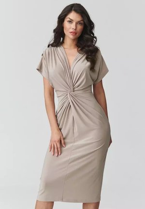 Kobieta z długimi ciemnymi włosami, ubrana w beżową sukienkę do kolan z marszczonym skrętem w talii, stojąca na tle jednolitego jasnego tła.