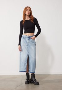 Jupe maxi en denim bleu clair avec fente à l'avant, présentant un ourlet effiloché. Assortie à un crop top à manches longues en maille noire et des bottes noires à lacets.