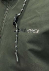 Olívazöld cipzáras dzseki, fekete-fehér mintás zsinórral és a mellkasán "Jack & Jones" logóval.