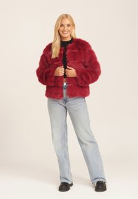 Red faux fur jacket with a boxy design and long sleeves, paired with light blue denim jeans and black chunky shoes.