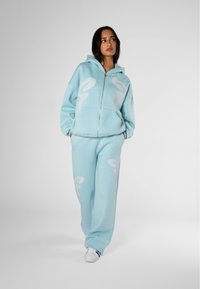 Felpa con zip color blu chiaro decorata con motivi astratti bianchi, abbinata a pantaloni della tuta coordinati. Realizzata in tessuto morbido, presenta tasche e cappuccio.