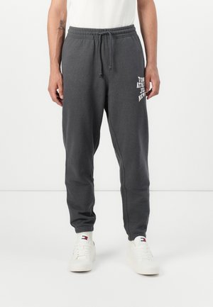 Homme portant un pantalon de jogging gris foncé avec un texte blanc sur la cuisse gauche, associé à des baskets blanches et des chaussettes blanches assorties sur fond blanc.
