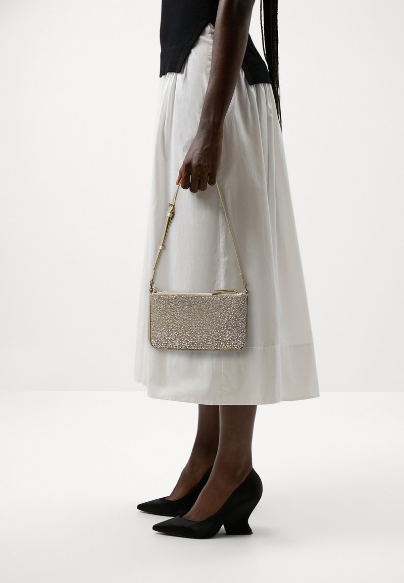 Personne portant une jupe midi blanche et un haut noir tient un petit sac à main beige texturé avec une bandoulière dorée, debout en talons noirs à bout pointu.