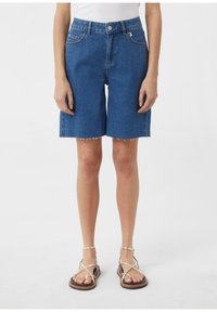 Bermudy dżinsowe w średnim niebieskim odcieniu, z prostym krojem, postrzępionym dołem i klasycznym pięciokomorowym wzornictwem. Połączone z sandałami na paski.