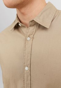Beige, long-sleeve shirt made of cotton blend; features a classic collar and white buttons along the placket, with a smooth texture.